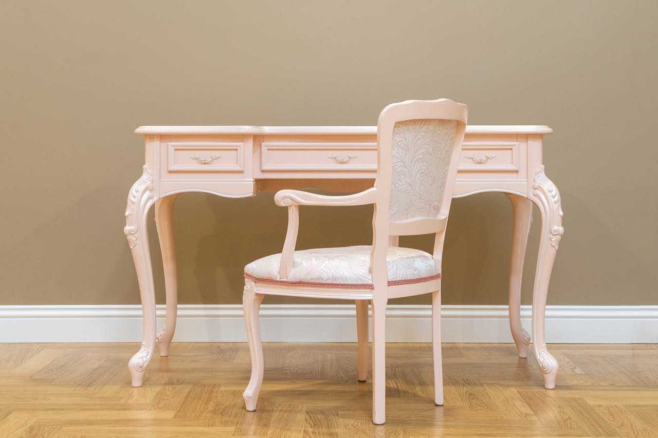 about-us A classic pink vintage desk and matching chair set on a wooden floor with beige wall, perfect for elegant interiors.