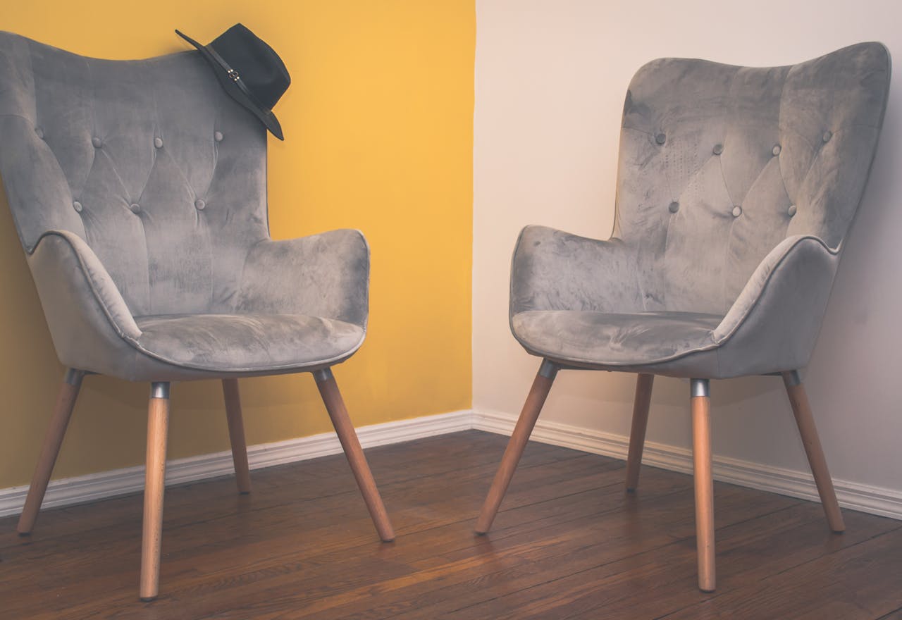 services-06 Two contemporary gray armchairs in a minimalist interior with a yellow accent wall and wooden flooring.